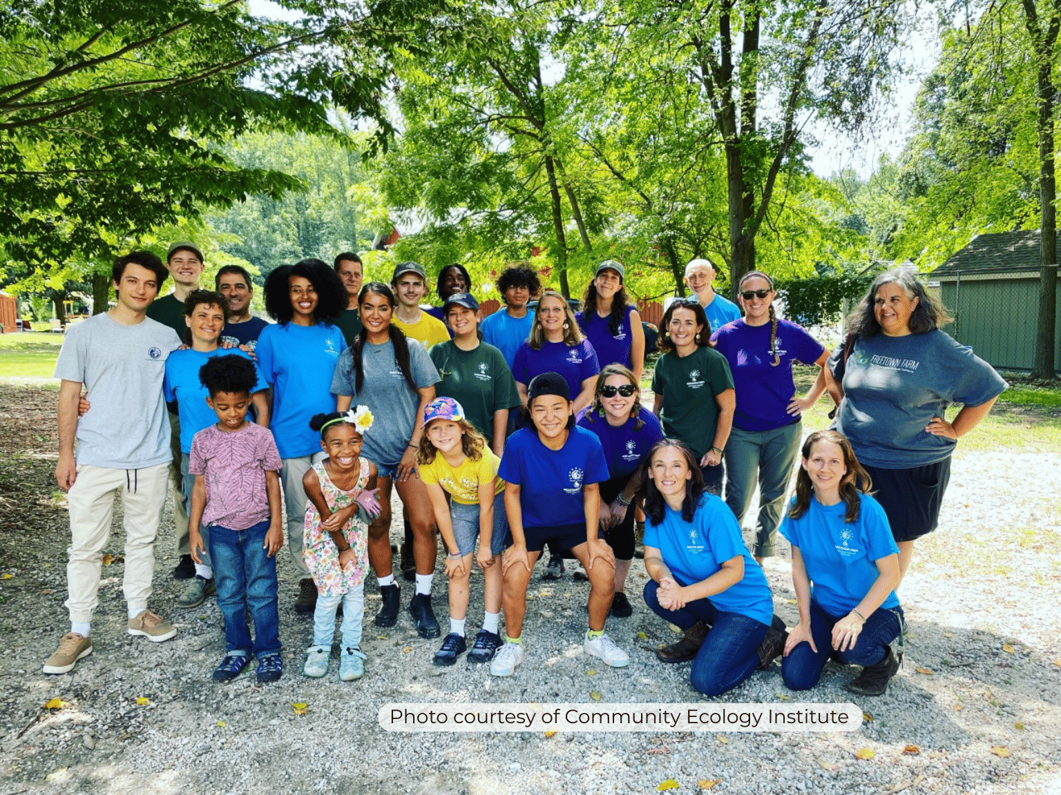 Group of adults and children standing on gravel with green trees in the background. Everyone is smiling at the camera.